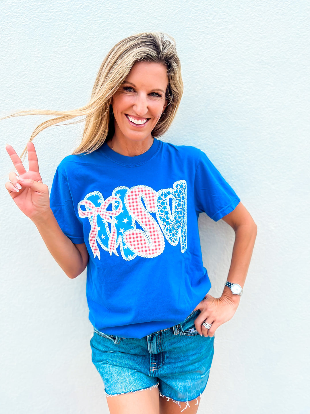 Woman wearing a blue Coquette USA graphic tee with gingham, lace, and bow design, paired with denim shorts and making a peace sign