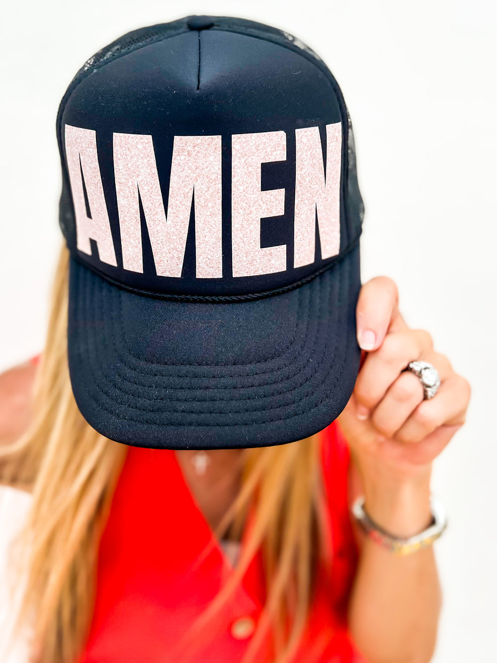 Woman wearing a black trucker hat with large rose gold glitter “AMEN” lettering, holding the brim down while smiling, styled with a red top