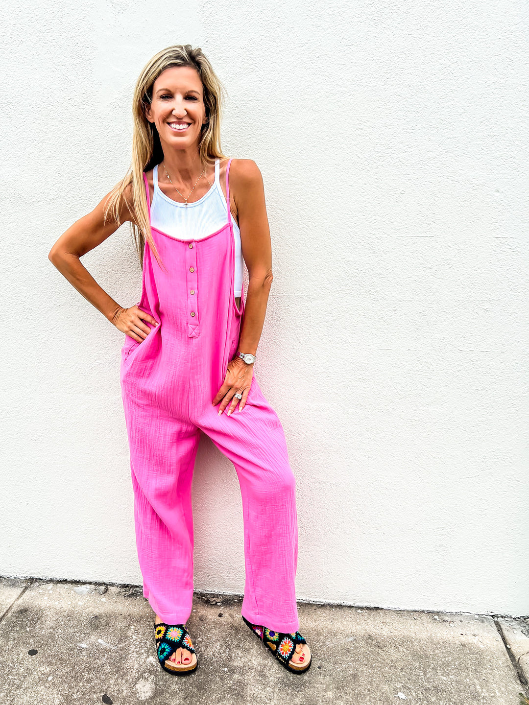 Woman wearing a pink gauze cotton jumpsuit with adjustable straps, front buttons, and wide legs, layered over a white tank top and smiling