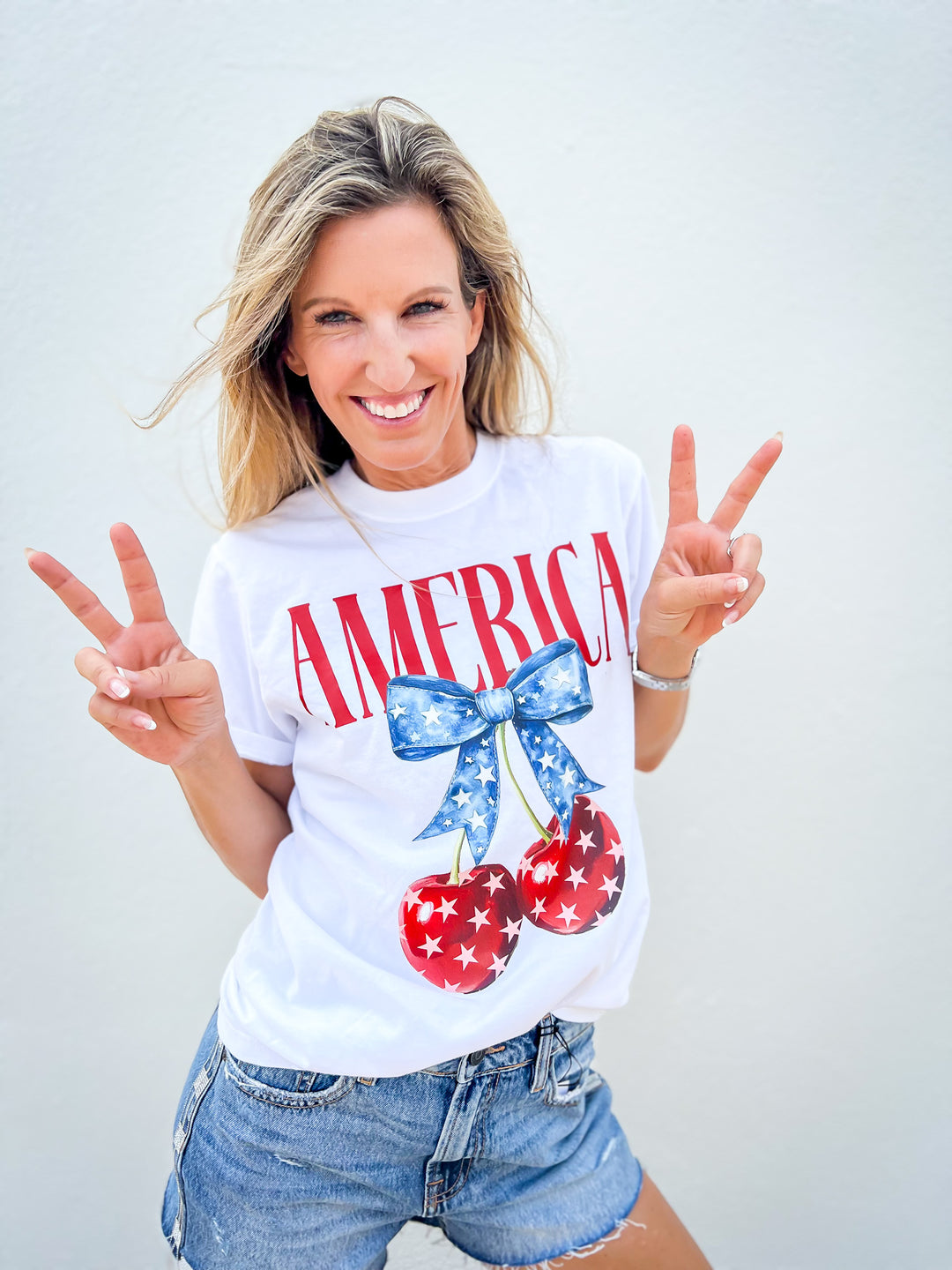 
Woman wearing a white Cherry Bomb graphic tee with red “America” text, patriotic cherry and bow design, and denim shorts against white background