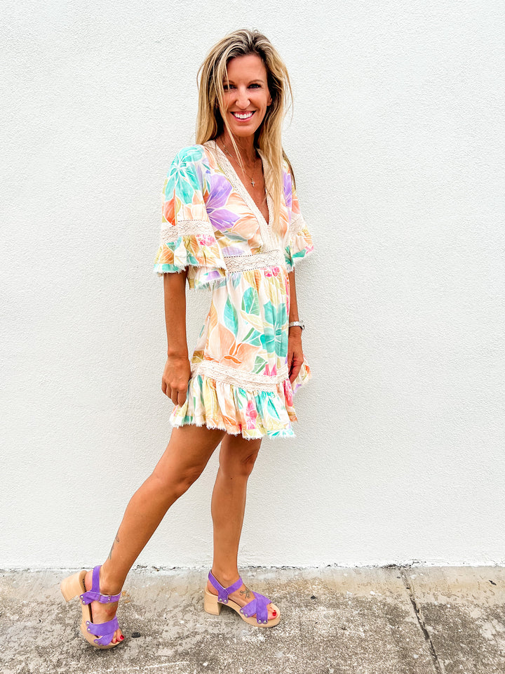 Woman wearing a colorful pastel floral mini dress with lace trim and ruffle hem, standing against a white wall and smiling
