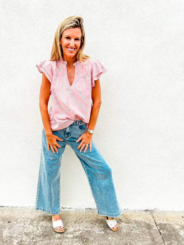 Woman wearing a red and white striped flutter sleeve top with a ruffle detail and notched neckline, styled with denim jeans, looking down and smiling