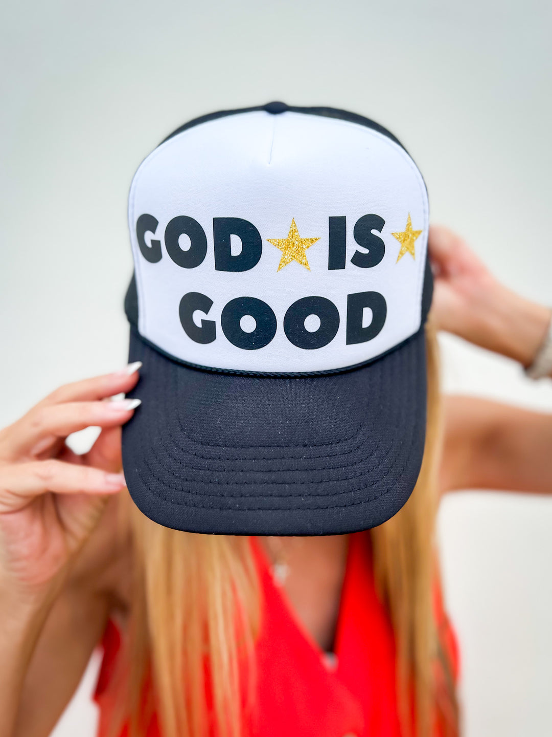 Woman wearing a black and white trucker hat with “God Is Good” text and gold stars, styled with a red sleeveless button dress

