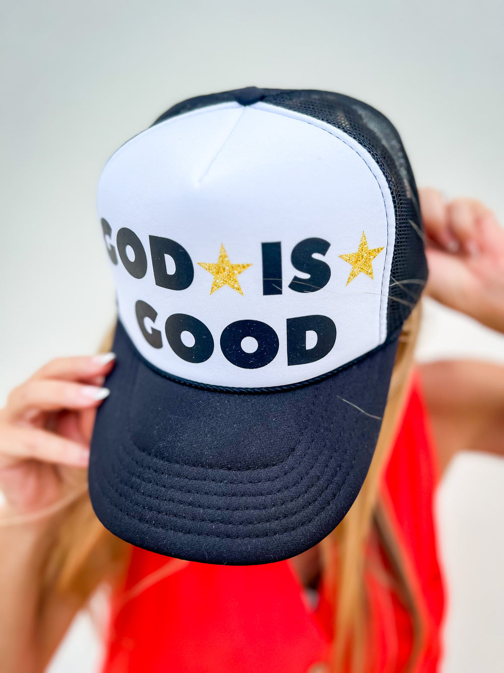 Woman wearing a black and white trucker hat with “God Is Good” text and gold stars, styled with a red sleeveless button dress

