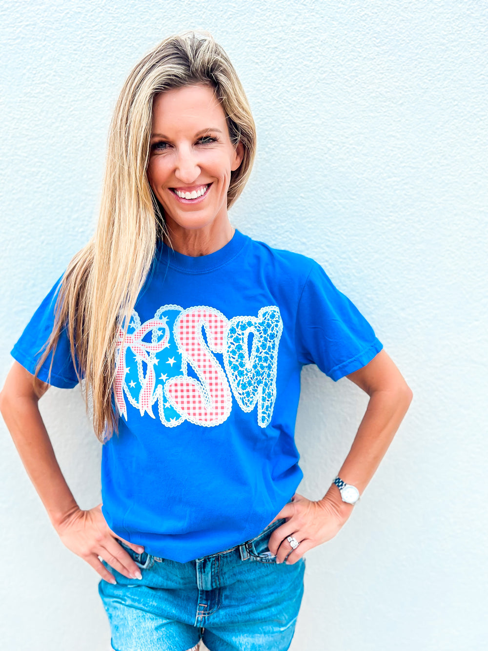 Woman wearing a blue Coquette USA graphic tee with gingham, lace, and bow design, paired with denim shorts and making a peace sign
