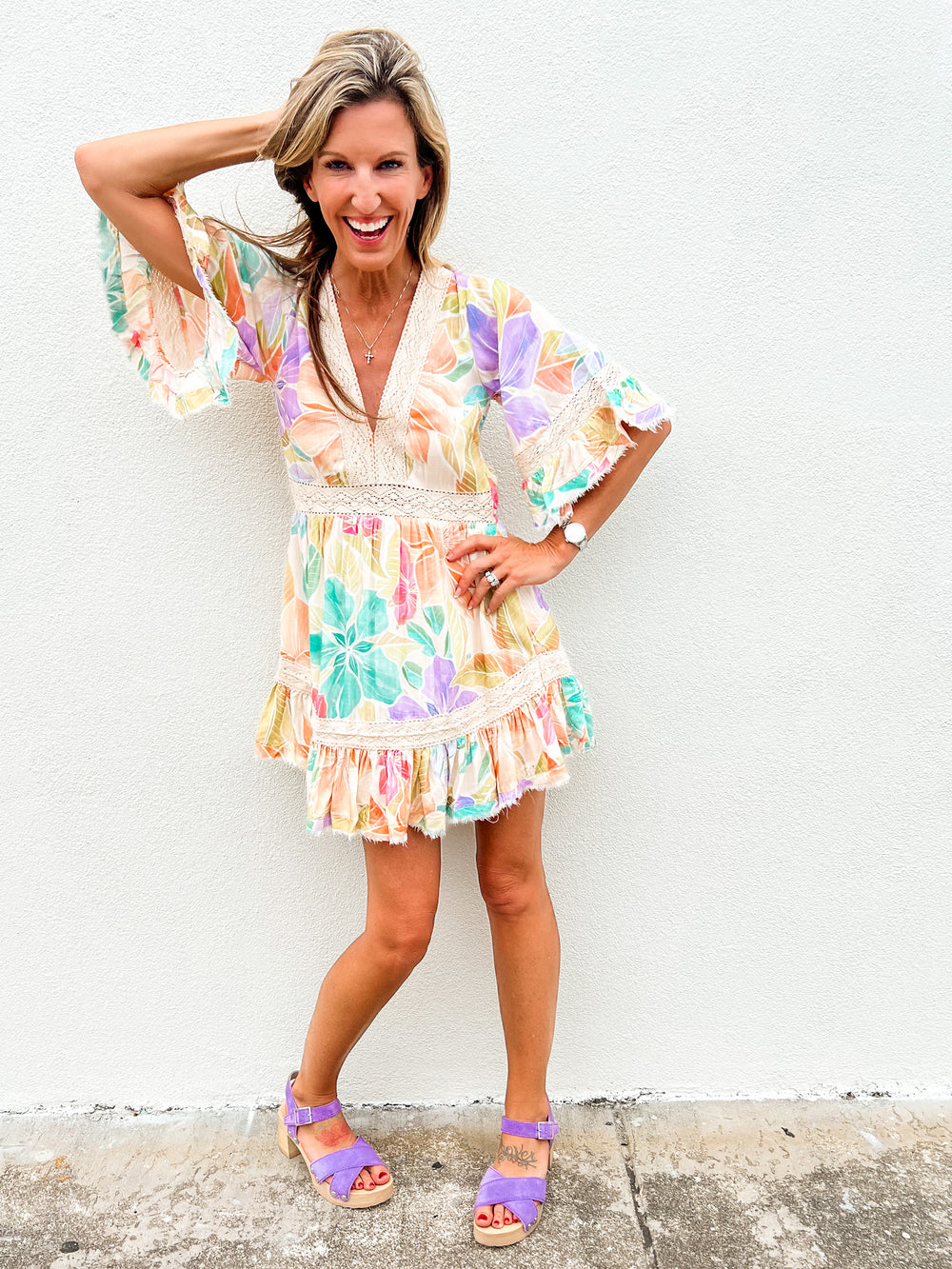 Woman wearing a colorful pastel floral mini dress with lace trim and ruffle hem, standing against a white wall and smiling