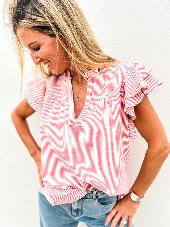 Woman wearing a red and white striped flutter sleeve top with a ruffle detail and notched neckline, styled with denim jeans, looking down and smiling