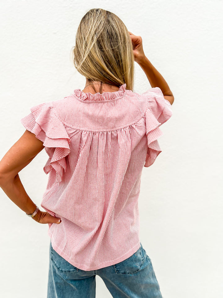 Woman wearing a red and white striped flutter sleeve top with a ruffle detail and notched neckline, styled with denim jeans, looking down and smiling