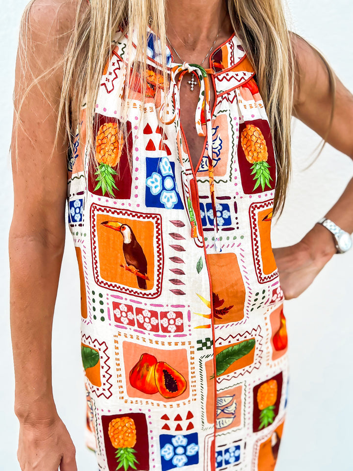 Woman wearing a sleeveless halter dress with a colorful tropical grid print featuring pineapples, toucans, and fruit designs, standing against a white wall