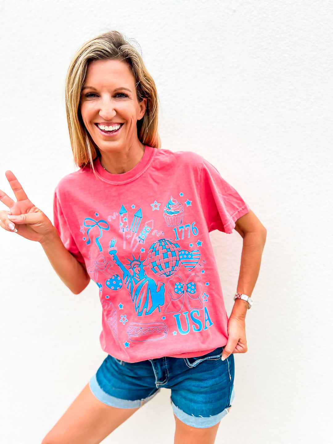 Woman wearing a watermelon red graphic tee with USA icons including the Statue of Liberty, hot dog, cupcake, disco ball, and stars