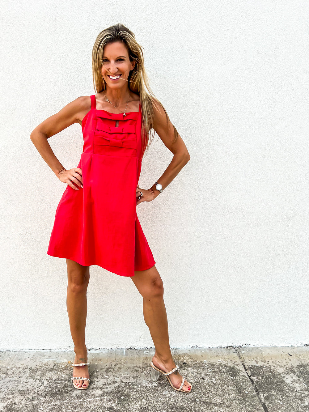 Woman wearing a red mini dress with tiered bow details on the chest, wide straps, and a flared silhouette, smiling in clear-heeled sandals