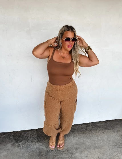 Woman wearing a brown tank top and pants against a white wall.