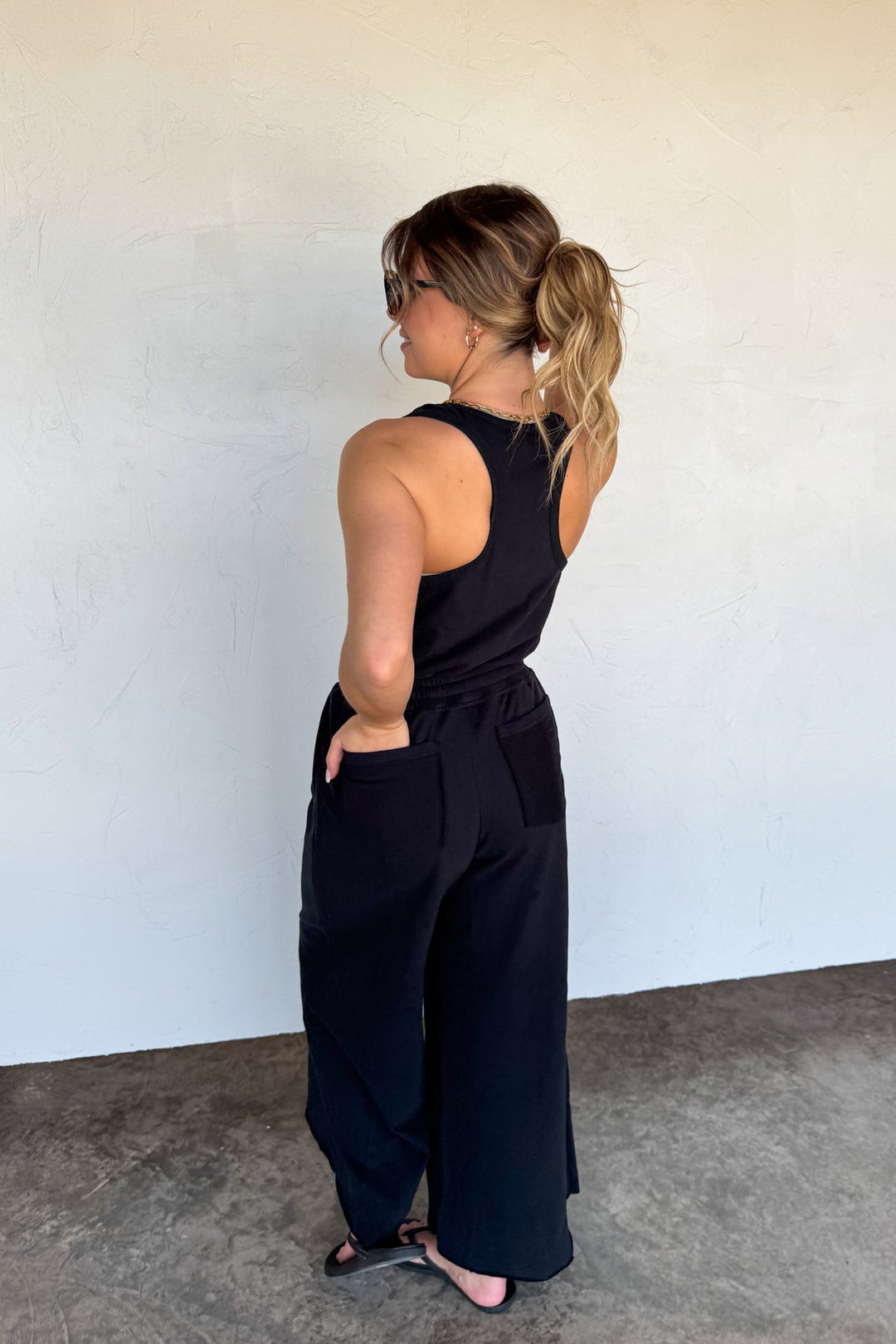 Woman wearing a black sleeveless top and wide-leg pants against a plain white wall.